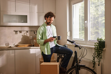A man stands on a box in his kitchen, holding a coffee mug and looking at his phone. Moving boxes...