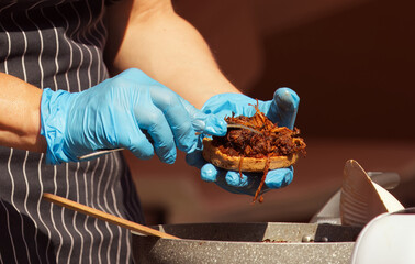 A professional chef wearing gloves carefully assembles a gourmet sandwich in a kitchen setting,...