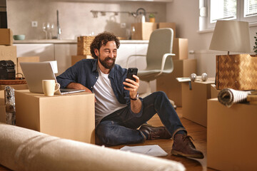 A man sits on the floor surrounded by moving boxes, smiling while using his smartphone. The cozy...