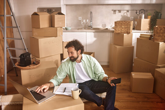 A man sits comfortably in a new home surrounded by unpacked boxes, using a laptop to organize tasks and holding a phone in his hand, as he adjusts to the new living space.