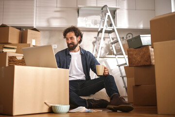 A man sits on the floor surrounded by unpacked boxes and a ladder, enjoying a coffee while working...