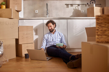 A man sits on the kitchen floor, focused on his laptop while holding a bowl. He is surrounded by...