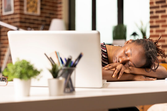 Overworked black businesswoman falls asleep at desk in home office after completing numerous tasks on laptop. Exhausted remote employee naps while seated, recovering from long hours of intense work.