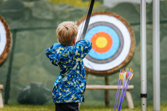 A young boy aims a bow at a colorful archery target, focusing intently while holding the string back, wearing a vibrant blue patterned jacket.