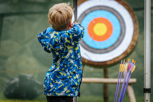 A young boy aims a bow at a colorful archery target, focusing intently while holding the string back, wearing a vibrant blue patterned jacket.