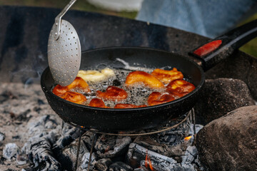 A black skillet with bubbling oil and golden pastries fries over hot coals, with a slotted spatula stirring the food in an outdoor rustic setting.