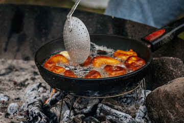A black skillet with bubbling oil and golden pastries fries over hot coals, with a slotted spatula stirring the food in an outdoor rustic setting.