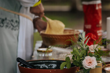 A person prepares batter in a woven bowl at an outdoor cooking setup, surrounded by rustic...