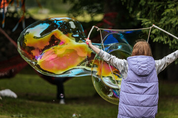 A child in a purple vest creates a large, colorful soap bubble using wands in a park setting,...