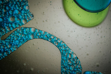 Vibrant green droplet encased in a blue fluid form, set against a neutral background with tiny suspended bubbles for added texture and depth.
