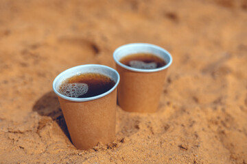 Two disposable coffee cups filled with frothy coffee resting in warm sand, creating a delightful...