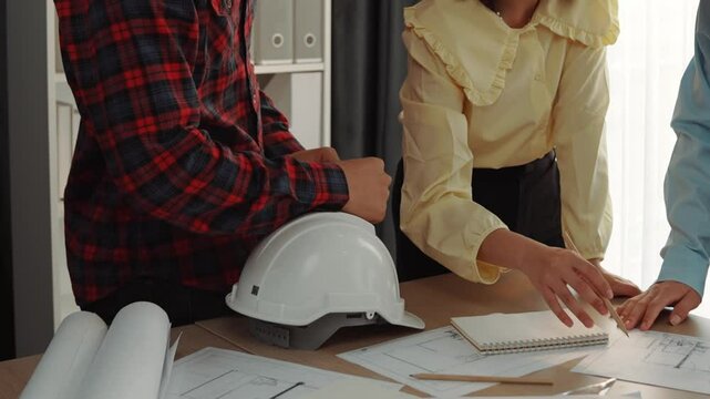 Smart architect and safety engineer discussion about building blueprint on table with architectural document, measurement tools and wooden block scatter around at modern office. Closeup. Delineation.