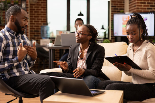 African american professionals discuss marketing strategies and business solutions, with female manager wearing eyeglasses providing updates to coworkers in cozy office lounge.