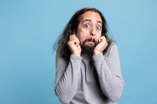 Joyful bearded man with long curly hair strikes a silly pose showing a carefree attitude. Playful behavior and comical face highlight his humorous personality in this studio against blue background.