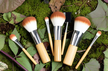 A collection of natural bamboo makeup brushes arranged on a bed of moss and leaves.