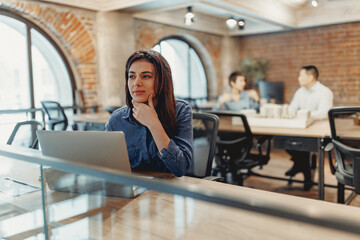 A focused woman works on her laptop in a stylish office while her colleagues discuss in the...