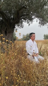 Mature master of karate in white kimono is sitting under the old olive tree and perfoming meditation, martial art concept