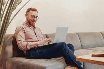 A joyful man deeply engaged with his laptop while sitting on a modern couch in a warm and inviting...