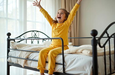 Obraz premium Joyful young girl in yellow pajamas on bunk bed, arms raised in excitement. Cozy bedroom interior, morning light streams in. Cheerful child, playful energy, happy expression, carefree childhood