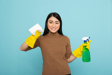 Asian housewife ready for action, holding detergent bottle and paper towel with cheerful smile....