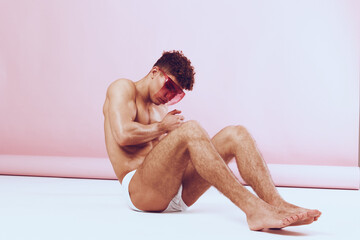 Young athletic man sitting on the floor, wearing pink sunglasses, showcasing a confident expression against a soft pink background. Fashion and self-expression concept.