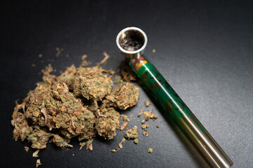 Presents green bong alongside a mound of cannabis, all set against a dark background. Selective focus