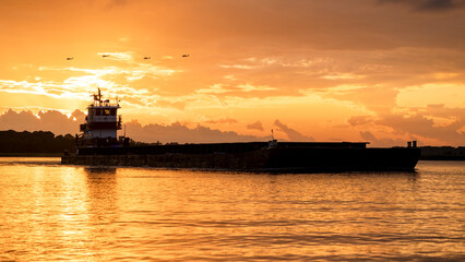 A barge going down the river at sunset with 4 helicopters silhouetted against the orange sky at Hilton Head and the gold sunset colors infusing everything