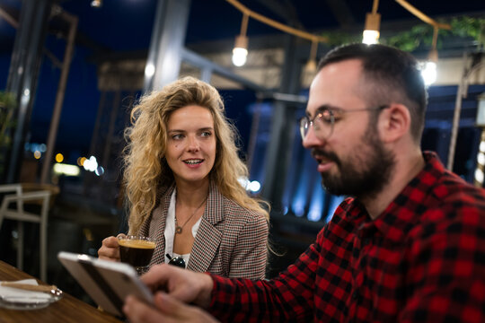 Friendly conversation between a woman and man in a cozy café setting during the night. They appear to be enjoying coffee together while discussing something on a digital device.