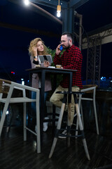 Casual business meeting at a rooftop bar with a man in plaid shirt using mobile phone and a woman with tablet both engaged in conversation under ambient lighting in an urban setting