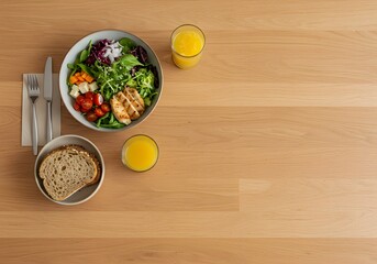 Healthy lunch of grilled chicken salad, bread, and orange juice, presented on a light wood table.