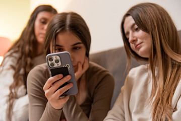Three young women sitting together on a couch, looking at a smartphone, engaging in social...