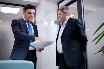Two professionals in a modern office discussing documents while dressed in formal business attire