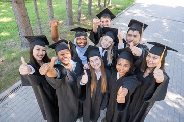 Students of different nationalities in graduation gowns give thumbs up. 