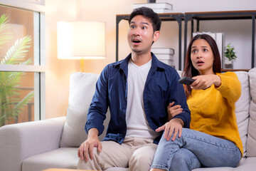 Young asian couple sitting on sofa in living room at home, reacting with surprise while watching...