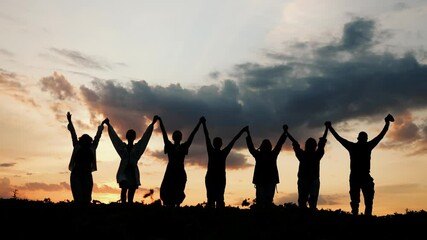 Silhouettes of a group of people, men and women, enjoying a country holiday. A group of seven people walk out into the sunset, holding hands and raising them up. - Powered by Adobe