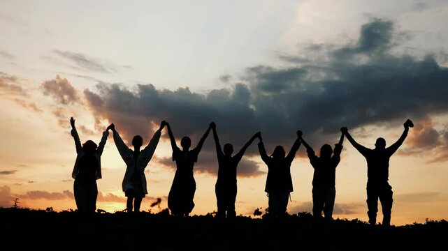 Silhouettes of a group of people, men and women, enjoying a country holiday. A group of seven people walk out into the sunset, holding hands and raising them up.