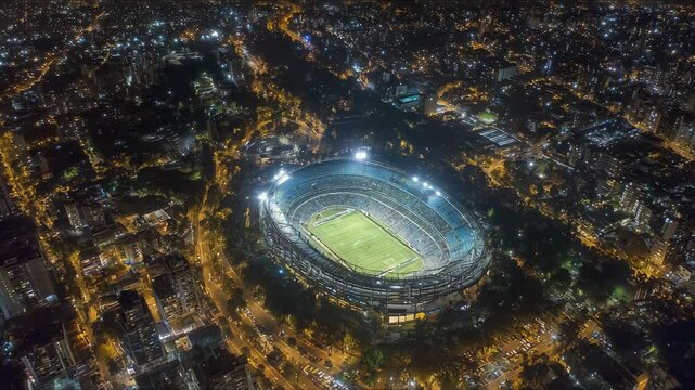 Nighttime aerial view of a football stadium glowing with bright floodlights, surrounded by city traffic and illuminated buildings