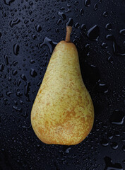 A green pear lies on a black background covered with water drops.
