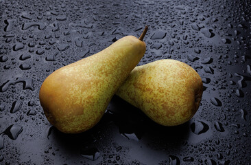 Two ripe green pears lie on a wet black surface covered with drops of water.