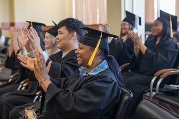Young people in graduation gowns listen to a speech and applaud. 