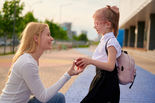 mom and daughter back-to-school moment. woman kneeling, handing red apple to smiling young girl with backpack. outdoor urban setting, warm sunlight, casual joyful scene.