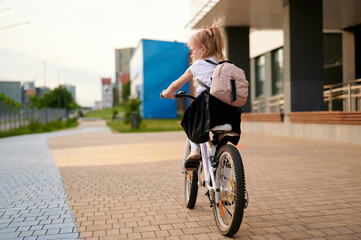 young girl riding bicycle with backpack on city sidewalk, embracing outdoor adventure. perfect for...