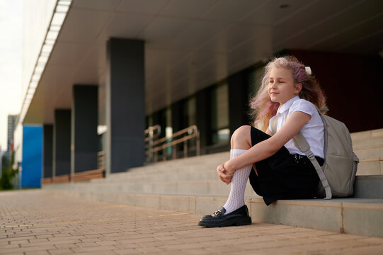 young girl with backpack sits on steps outside modern building, enjoying calm morning light. wearing school uniform, she appears thoughtful, capturing essence of school life