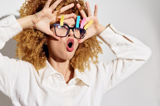 Creative woman making funny faces while holding colorful sticky notes on glasses, showcasing playful expression, studio background Playful, expressive, joyful emotions
