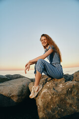 smiling woman sitting on rocks by the sea wearing casual blue outfit and sneakers during sunset with clear sky and warm light