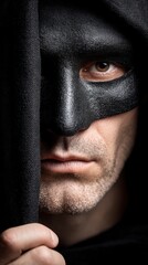 Close up portrait of a man wearing a textured black mask and dark hood revealing one eye