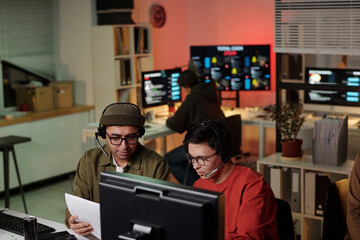 Two young adult men wearing headsets sitting at desk discussing script while working on computer in...