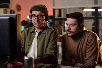 Two young adult men, one South Asian and one Caucasian, wearing headsets sitting closely at...