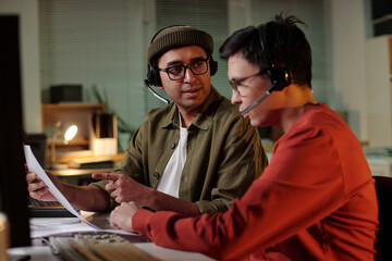 Biracial man wearing headset instructing Indian man with headset while reviewing documents at...