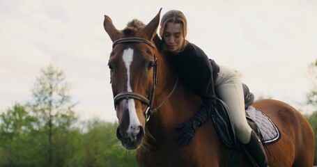 An elegant young woman smiles at the camera while lying on a horse and stroking its muzzle